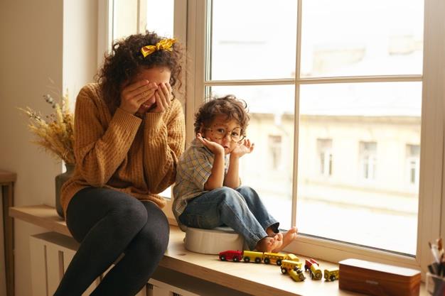 Cozy scene of happy family indoors. attractive young female with curly hair enjoying sweet moments of maternity, sitting in large windowsill, playing seek and hide with adorable infant child Free Photo