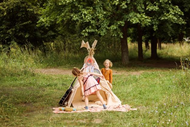 Funny girls are playing next to the wigwam. kids are dressed in boho or hippie style and play hide and seek. summertime, outdoors in the forest or park. family weekend. Premium Photo